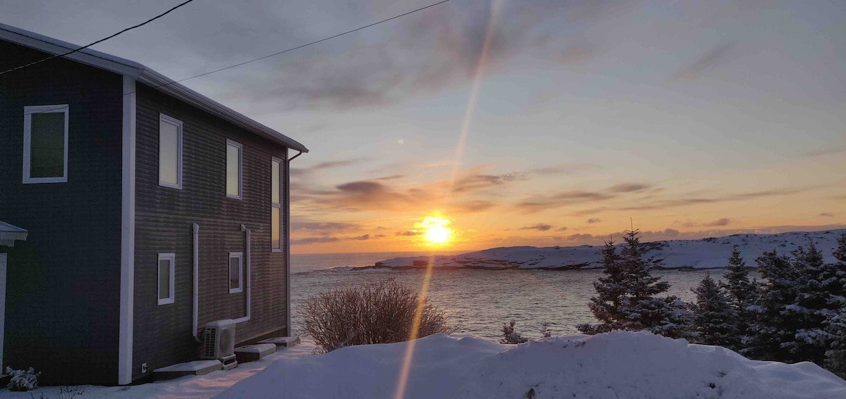 The exterior view captures a snow-covered landscape, with a modern two-story home positioned to the left. A vibrant sunset casts an array of warm colors across the sky, illuminating the ocean in the background, while silhouettes of trees are visible in the foreground.
