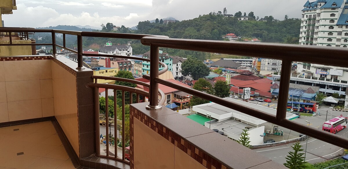 A balcony railing curves gracefully along the edge, offering views of the surrounding hills and nearby structures. The landscape features a mix of buildings and greenery under a cloudy sky, with glimpses of colorful rooftops and parked vehicles below.