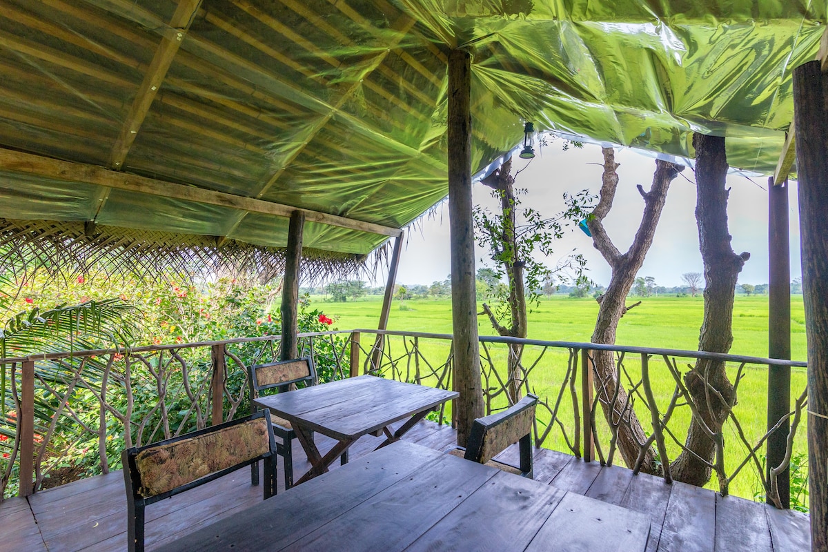 A rustic outdoor seating area features a wooden table surrounded by benches, set under a thatched roof. Lush greenery and vibrant flowers are visible in the surrounding landscape, with expansive views of a serene rice field stretching into the distance.