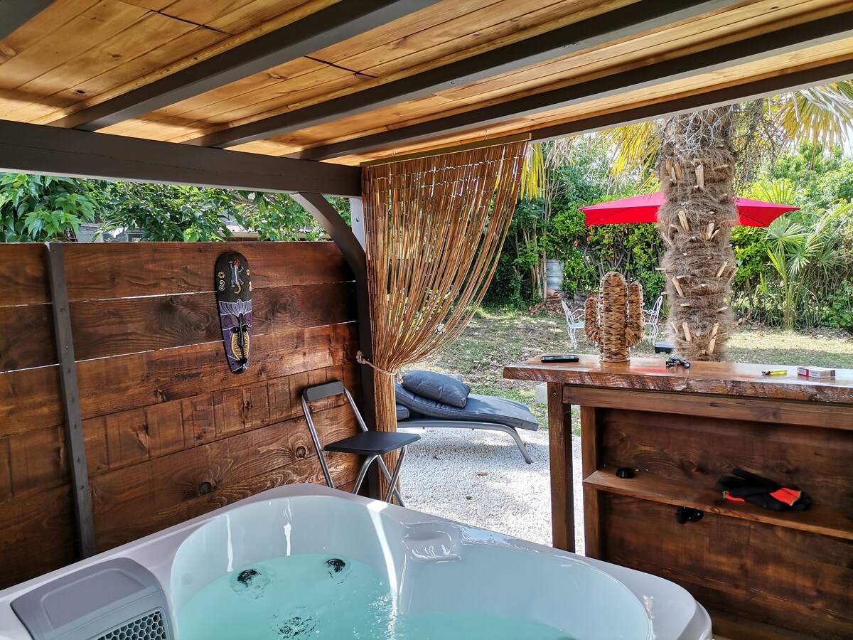 A private jacuzzi is positioned under a wooden roof, surrounded by lush greenery. A chair and a rustic bar are visible, with a sun lounger and an umbrella in the background. Natural light filters through the slatted design, creating a serene relaxation space.
