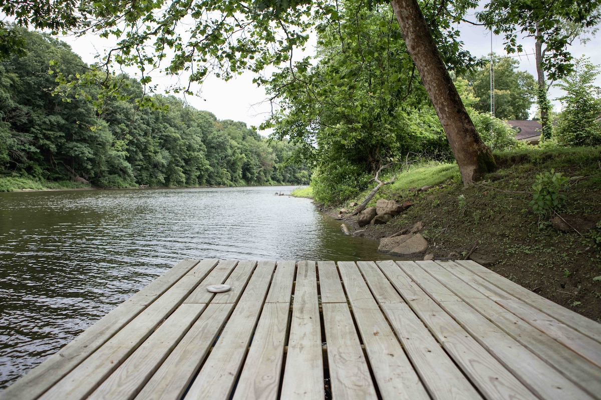 A wooden dock extends over calm waters, surrounded by lush greenery. The shoreline features gentle slopes and stones, while tall trees frame the view, creating a serene and calming atmosphere along the water's edge.