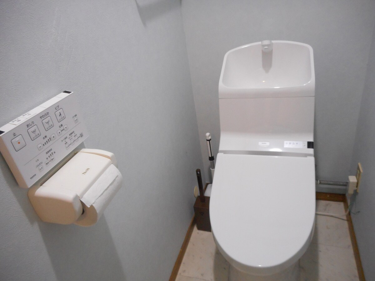 A modern bathroom is featured, showcasing a sleek toilet with integrated controls on the wall beside it. A roll of toilet paper is conveniently located nearby, and cleaning supplies are discreetly stored in a holder on the floor.