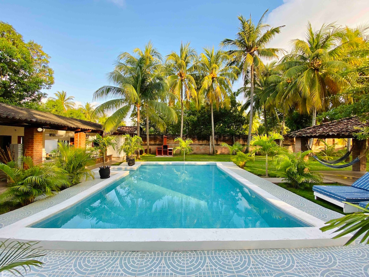 The rectangular pool is surrounded by palm trees and a well-maintained lawn. Comfortable lounge chairs and hammocks are positioned nearby for relaxation. Pavilions and tropical plants enhance the serene atmosphere, creating a peaceful outdoor retreat.