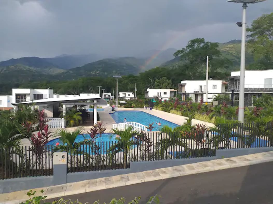 The image showcases a community area featuring multiple swimming pools, surrounded by lush greenery and trees. Mountains are visible in the background, and a faint rainbow arcs across the sky. The scene captures a tranquil setting intended for relaxation and recreation.
