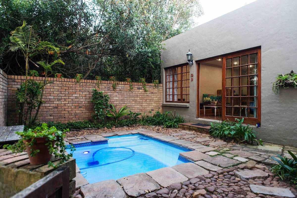 A private pool is framed by stone tiles and surrounded by lush greenery. The cottage entrance features large glass doors, allowing natural light to fill the space. A stone wall provides privacy, with a few potted plants adding to the serene garden setting.