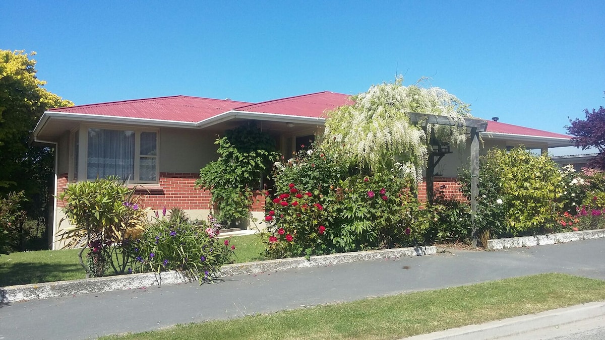 A large house with a red roof is surrounded by vibrant flowering shrubs and greenery. A well-maintained garden leads up to the entrance, with clear blue skies overhead. The home features windows with light curtains, inviting a sense of openness and connection to the outdoors.
