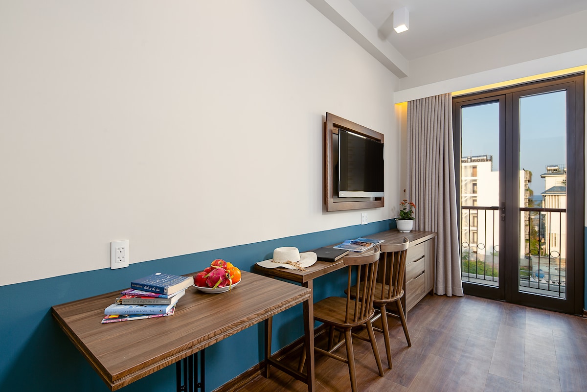 A modern studio features a wooden dining table and chairs, positioned beside a large window that allows natural light to enter. A television is mounted on the wall across from the seating area. Colorful books are neatly arranged on the table, complemented by a bowl of fresh fruit.