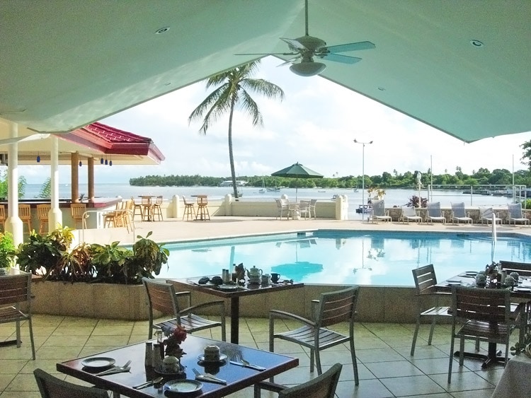 A dining area is set beside a pool with clear blue water, featuring a shaded patio and tropical plants. Tables are neatly arranged, and a view of the beach and palm trees is visible beyond the pool area, creating a serene atmosphere.