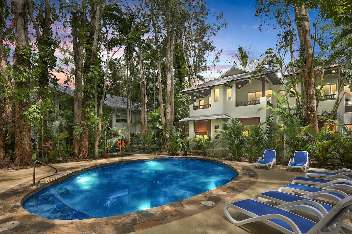 A tranquil swimming pool is framed by lush tropical landscaping and towering trees, creating a serene environment. Comfortable lounge chairs line the edge of the pool, inviting relaxation under the natural canopy. Soft evening light casts a warm glow over the surroundings.