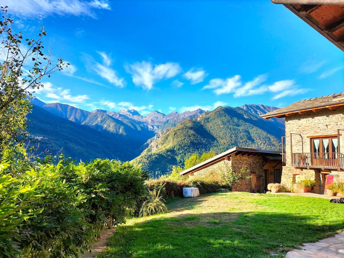 A scenic view of rolling mountains is displayed under a clear blue sky, with scattered clouds. The well-maintained garden features lush greenery, while a charming stone building is visible on the right side, enhancing the serene rural setting.
