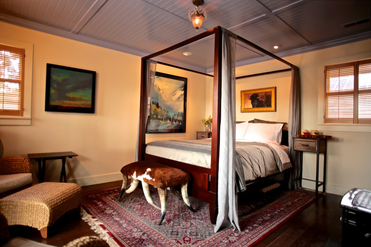 A cozy bedroom features a four-poster bed with a light-colored bedding set. A decorative ottoman with a cowhide pattern sits at the foot of the bed. Artwork adorns the walls, and natural light filters through the wooden blinds of the windows.