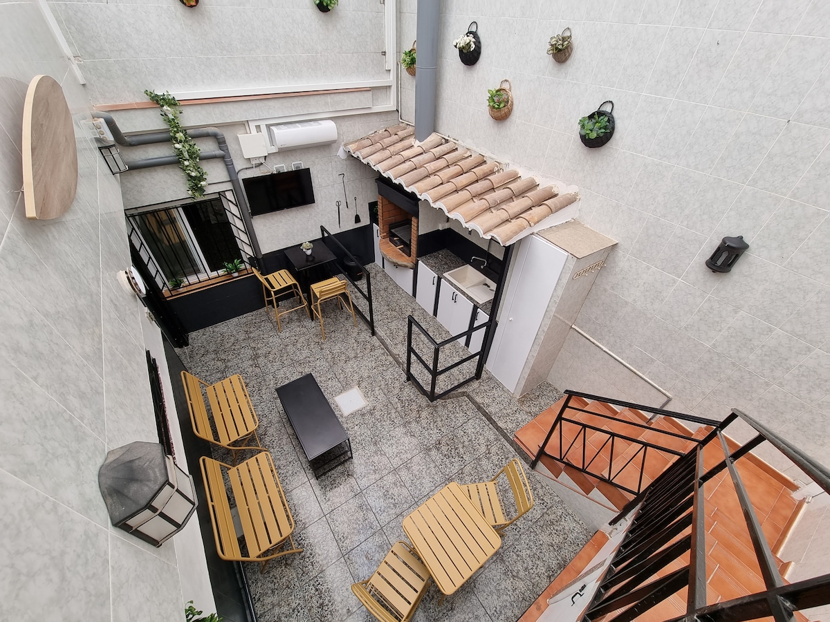 An inviting private patio is equipped with wooden lounge chairs and a central coffee table. A barbecue area is visible, complemented by built-in storage. Vertical planters with greenery add a touch of nature, and the surrounding walls are tiled, providing a clean atmosphere.