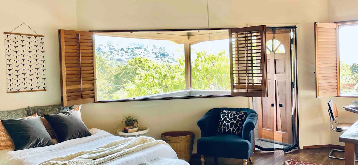 A spacious studio features a large window with wooden shutters, framing views of the surrounding greenery. The cozy bed is adorned with neutral linens. A comfortable chair and a small table are positioned nearby, emphasizing the room's relaxed ambiance.