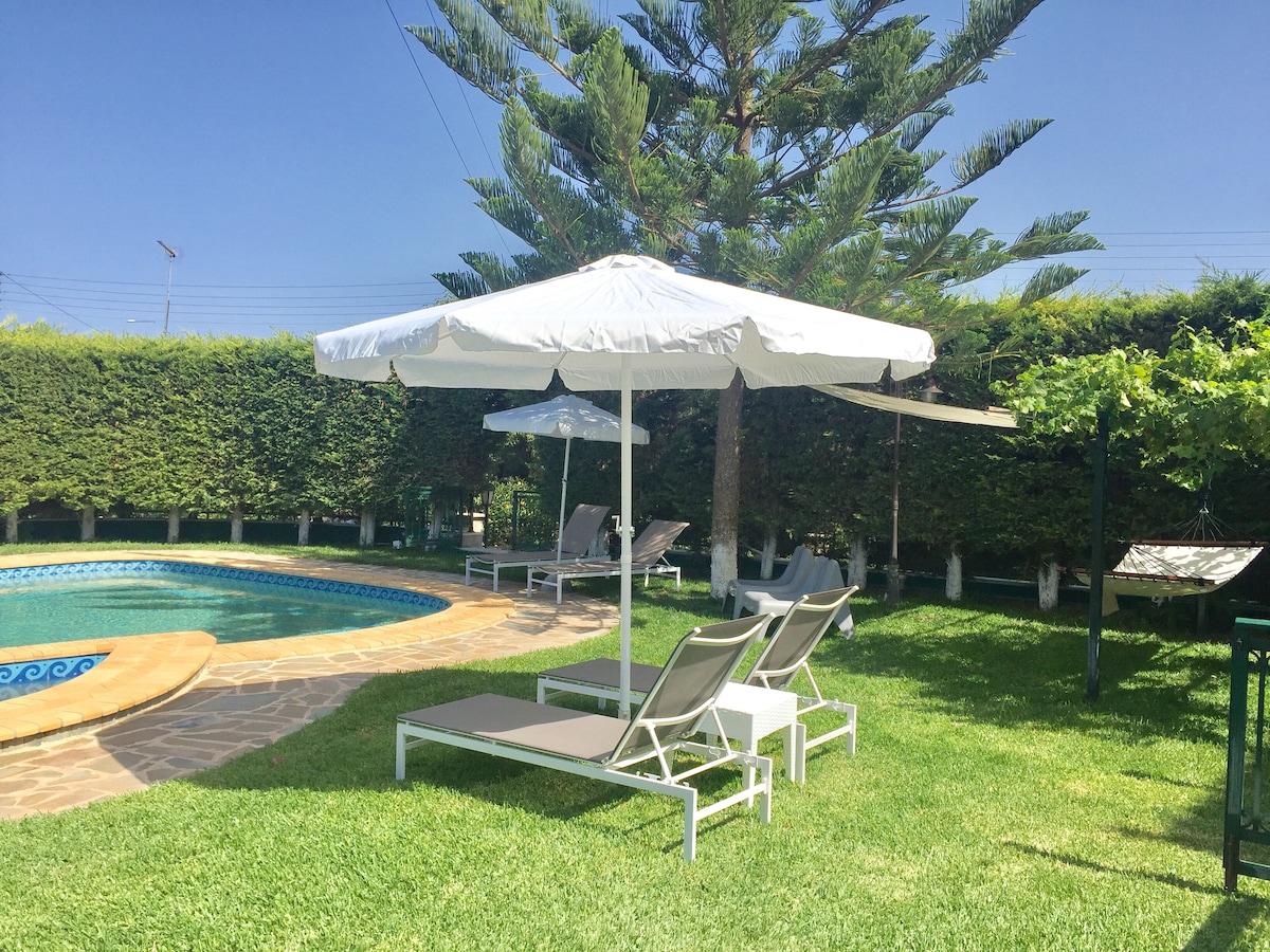 A serene outdoor space features a private swimming pool surrounded by lush greenery. Two lounge chairs sit beneath a white umbrella, offering shade. Nearby, a few sun loungers and a tree provide additional relaxation areas, creating an inviting atmosphere for outdoor enjoyment.