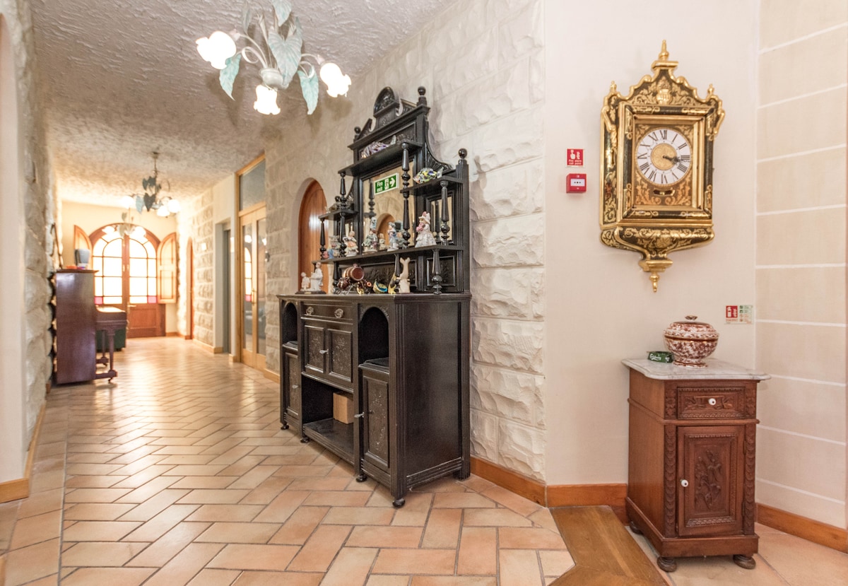 A long hallway is adorned with textured stone walls, featuring a wooden console table displaying decorative items. An ornate wall clock and a small cabinet add to the decor, while natural light filters through arched windows at the far end.