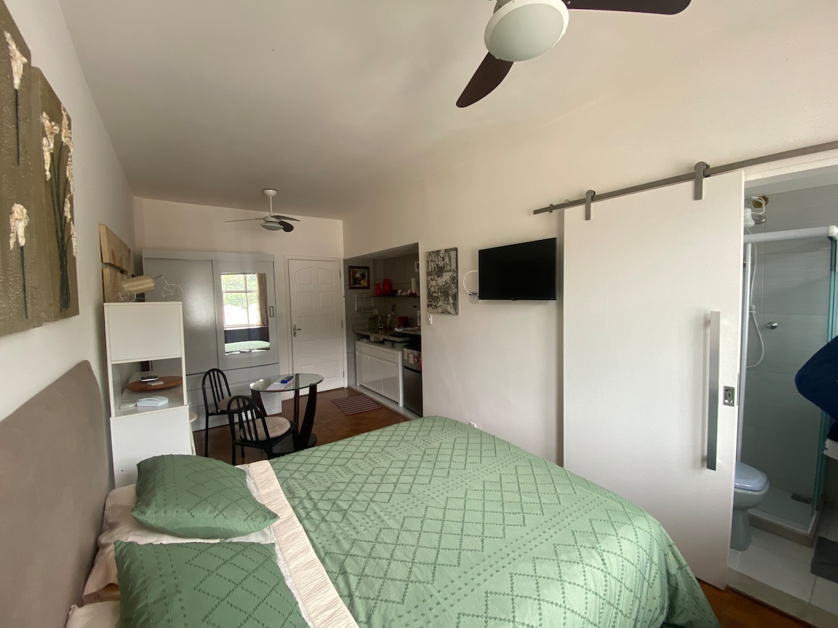 A cozy studio is showcased, featuring a spacious bed with a textured green blanket. A sliding barn door leads to a modern bathroom, while a small kitchen area with a dining table is visible. Natural light fills the space from the window.