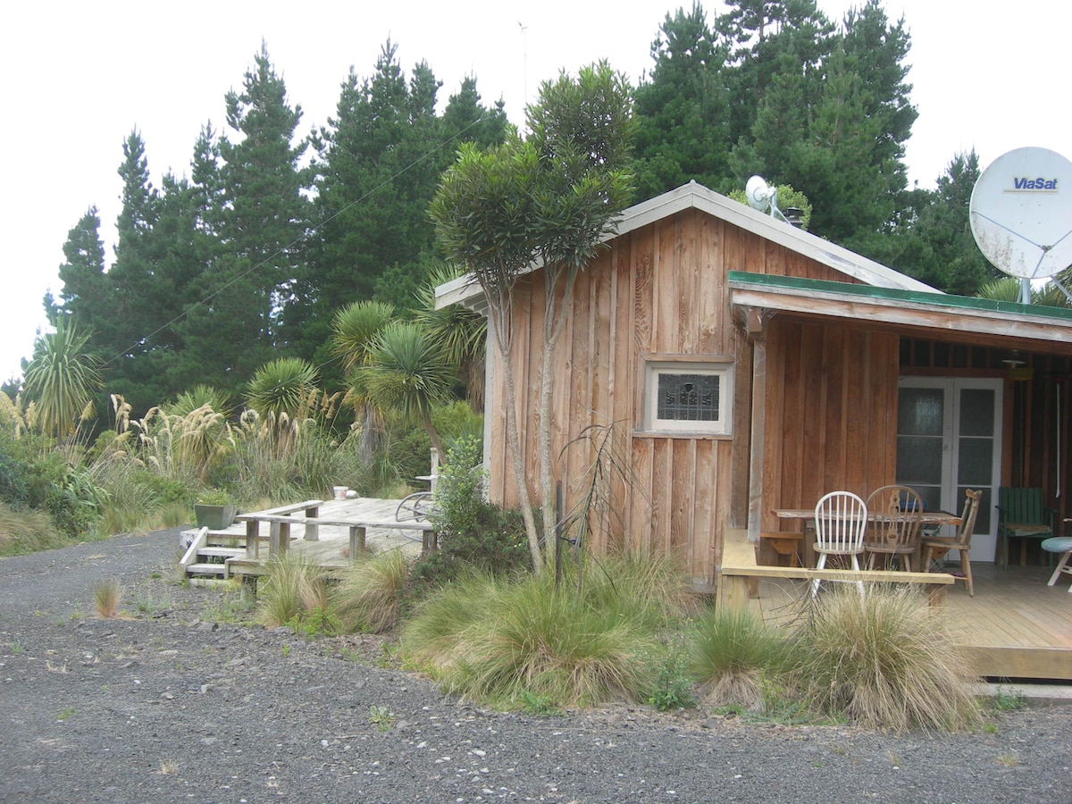 The cottage, constructed with natural wood, is set among native vegetation and surrounded by tall trees. A spacious deck is visible, featuring a table and chairs, inviting relaxation in the serene outdoor setting. Access to the cottage is provided by a gravel pathway.