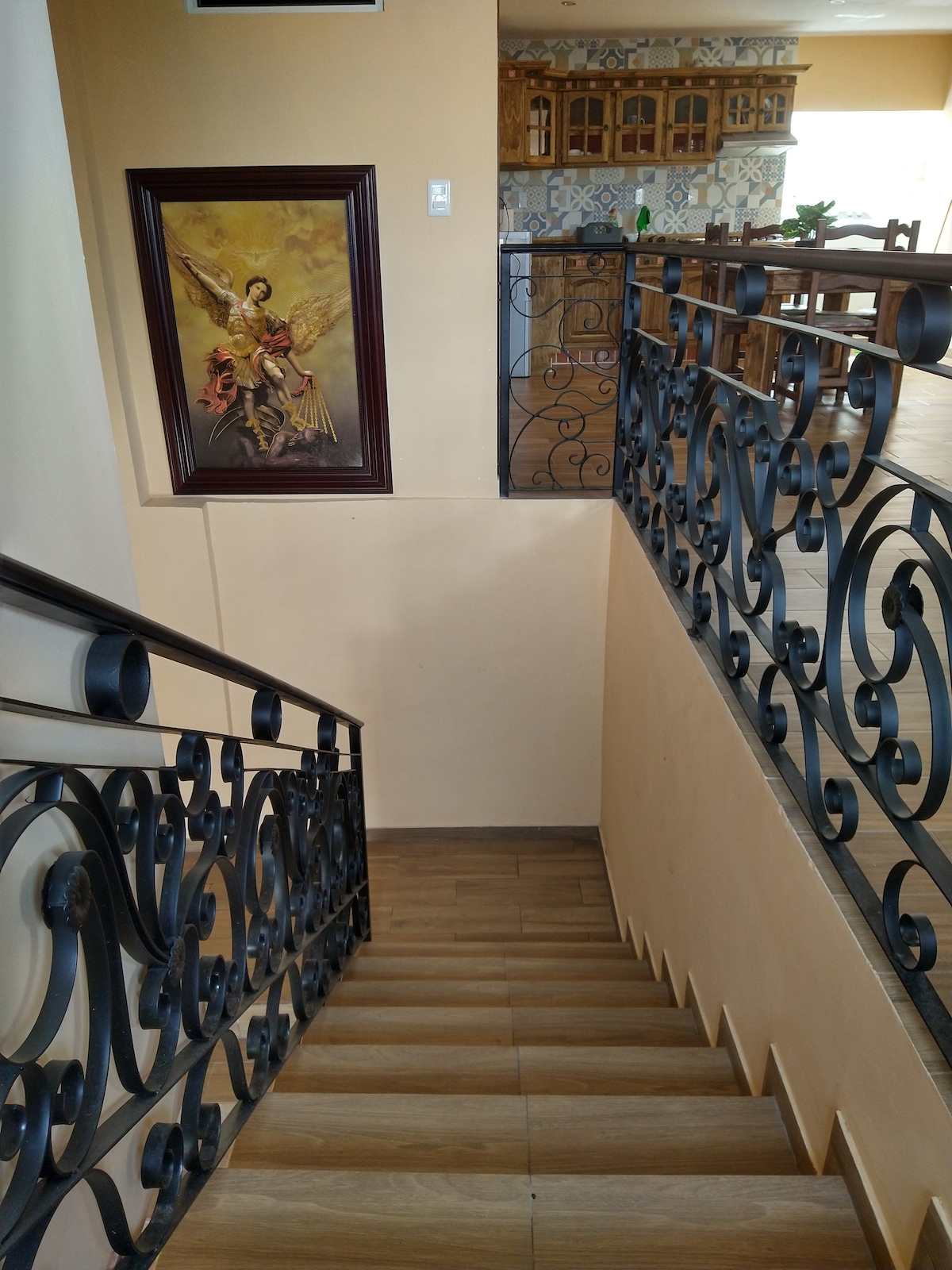 A well-lit staircase features smooth wooden steps and an elegant wrought-iron railing, leading down to a spacious area. A framed artwork can be seen on the wall, adding a touch of artistic detail to the environment.