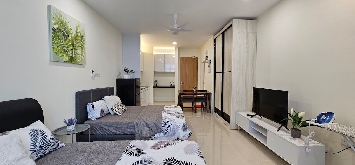 A modern studio unit features two spacious queen beds with decorative throw pillows. A television is positioned on a white entertainment unit near a dining table with chairs. A fully equipped kitchen is visible in the background, along with natural light sourced from a window.