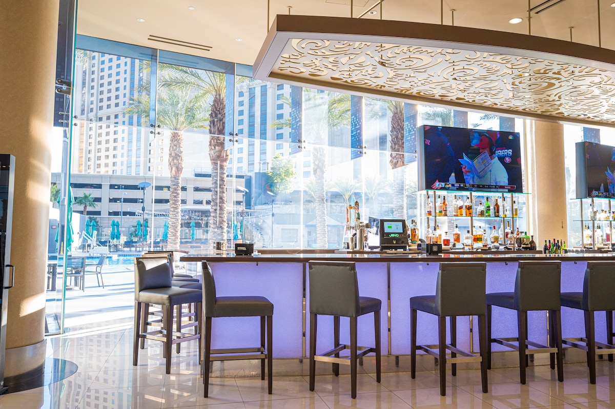 The image features a stylish bar area with high-backed chairs arranged around a curved counter. Large glass windows allow natural light to fill the space, offering a view of palm trees and an outdoor pool area. A bar shelf displays various bottles and a television is visible in the background.