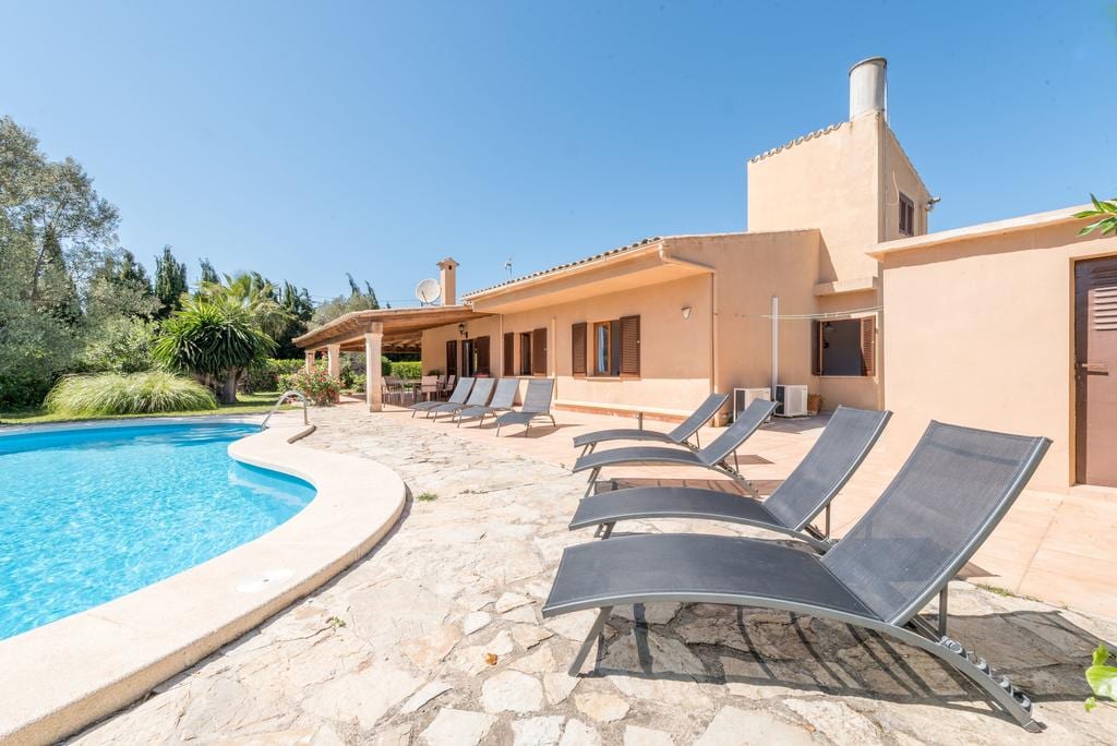 A sunlit terrace surrounds a swimming pool, lined with several loungers for relaxation. The villa features a warm exterior with large overhangs, complemented by lush greenery in the background. Clear skies enhance the inviting outdoor space, ideal for leisure and sunbathing.