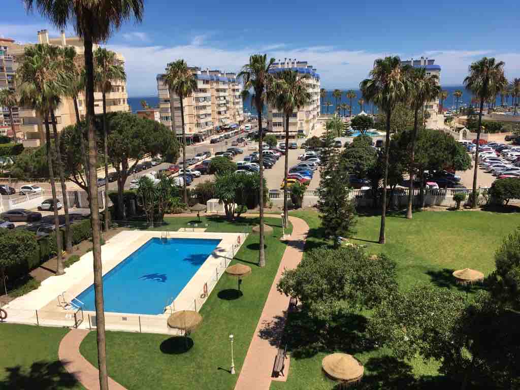 A view from above showcases a spacious outdoor area featuring a blue swimming pool surrounded by palm trees. Adjacent are well-maintained green lawns and a walking path. The distant view highlights the beachfront and neighboring buildings, creating a relaxing atmosphere.