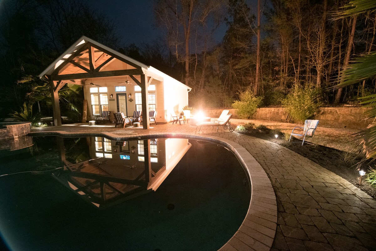 A tranquil pool house is illuminated at night, reflecting on the calm waters of the nearby pool. Gentle landscape lighting highlights the stone pathway and provides a warm atmosphere, while seating areas around a fire pit invite relaxation amid the surrounding trees.