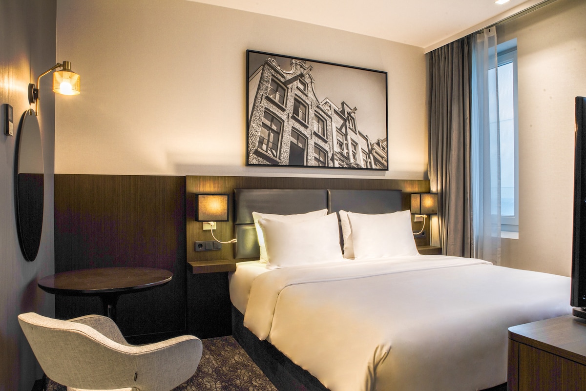 A spacious Standard Studio Suite features a large bed with crisp white linens, complemented by a round table and a stylish chair. A black-and-white cityscape photograph adorns the wall, and natural light filters in through a large window, creating a serene atmosphere.