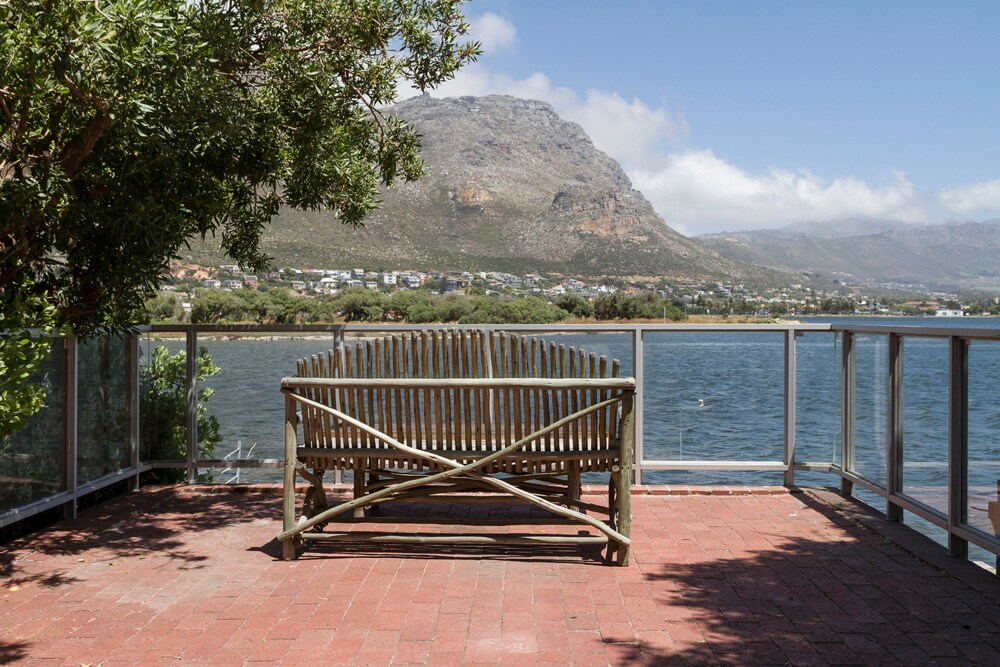 A wooden bench is positioned on a brick patio, overlooking a calm body of water. The scenery includes a distant mountain and lush greenery framing the view. Clear skies and soft lighting enhance the tranquil atmosphere of the outdoor space.
