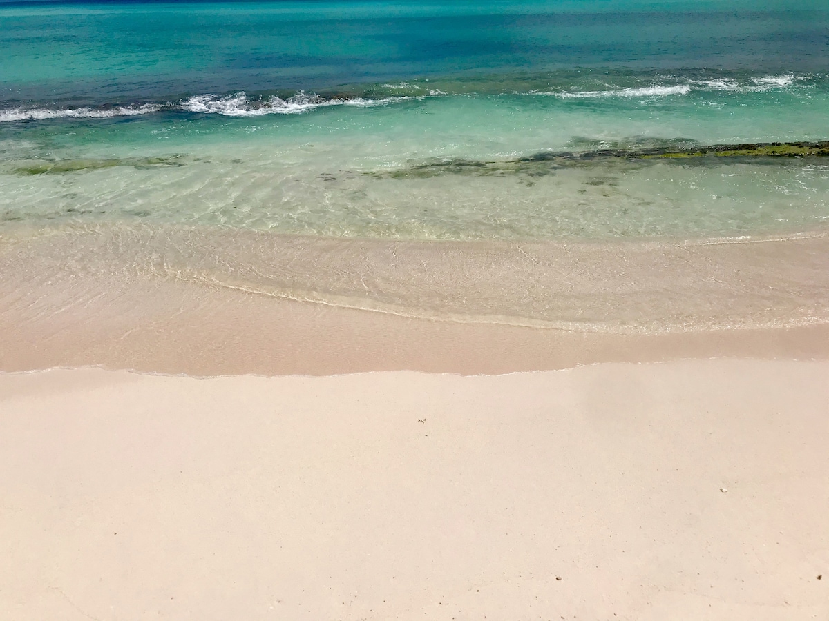 Soft sandy beach stretches across the foreground, meeting the calm turquoise waters at the shoreline. Gentle waves create ripples in the water, while the clear sky reflects the tranquil scene on the beach.