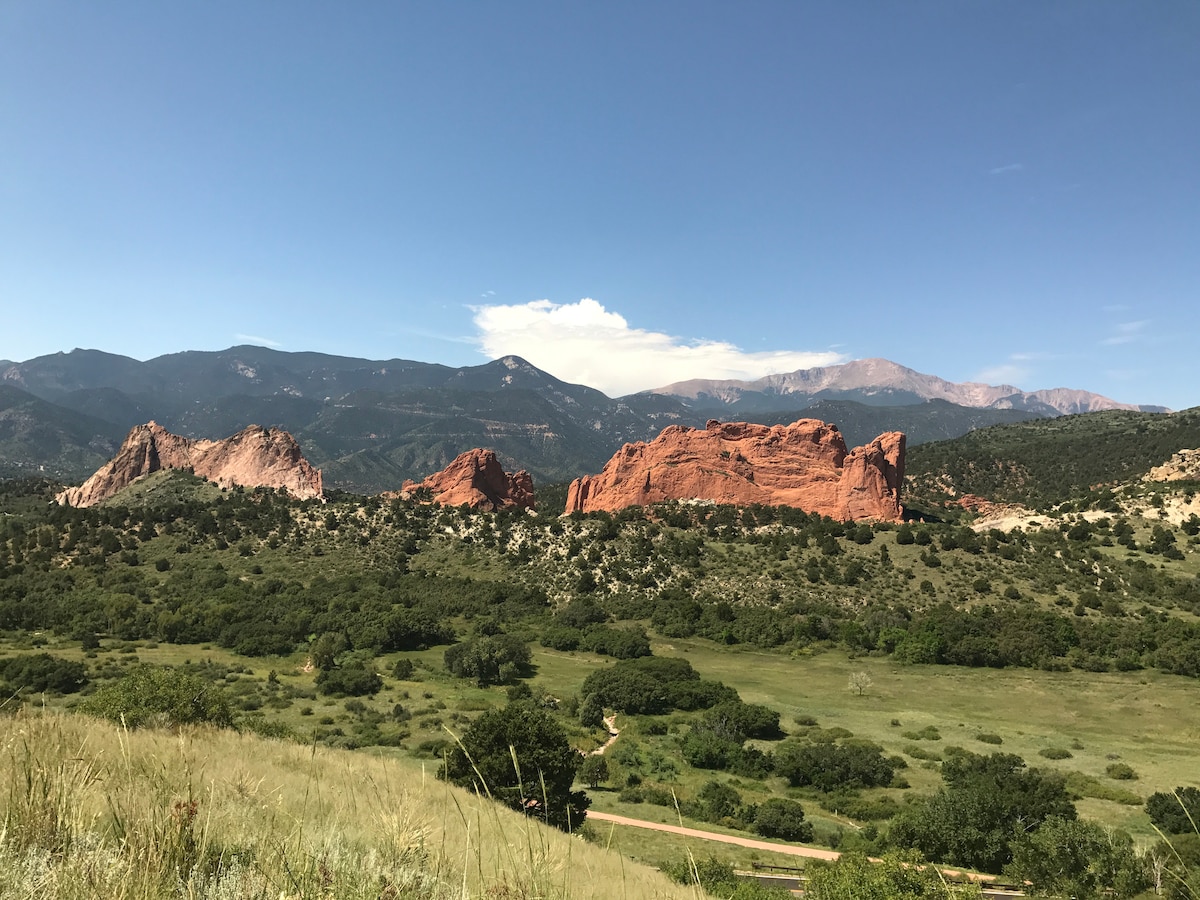 A panoramic view displays the striking red rock formations of Garden of the Gods, set against a backdrop of green hills and distant mountains. The sky is clear with a few clouds, offering a sense of openness and natural beauty.