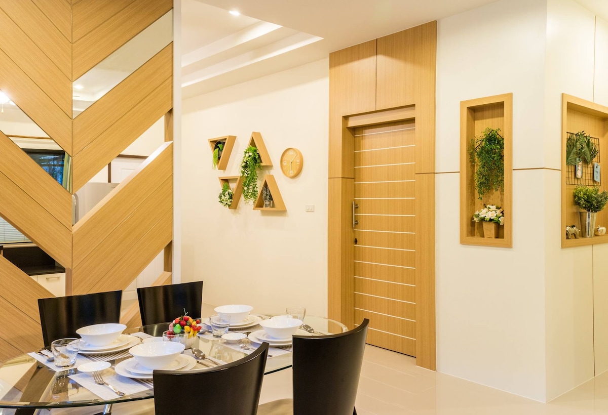 A modern dining area is seen with a round glass table set for four, featuring elegant dinnerware. Decorative wall shelves with plants add a touch of greenery. The entrance door is situated nearby, framed by light neutral walls with wooden accents, creating a stylish and inviting space.