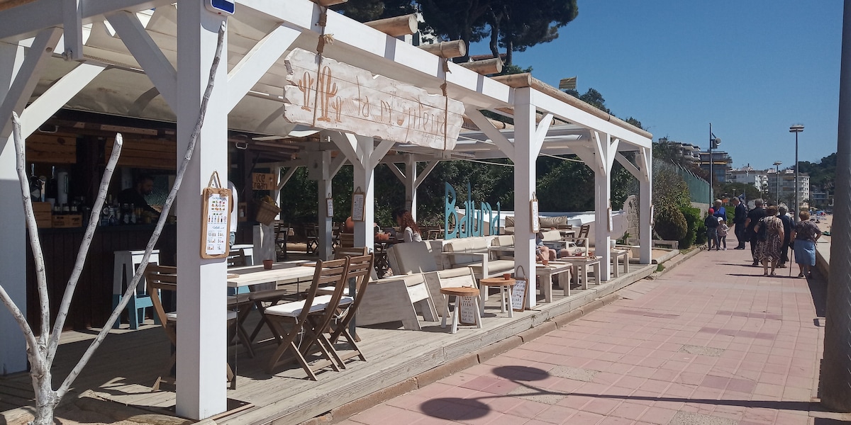An open-air dining area is featured with white wooden beams supporting a shaded roof. Several wooden tables and chairs are arranged for seating. A banner is displayed overhead, and the surrounding path is lined with lively visitors enjoying the day.