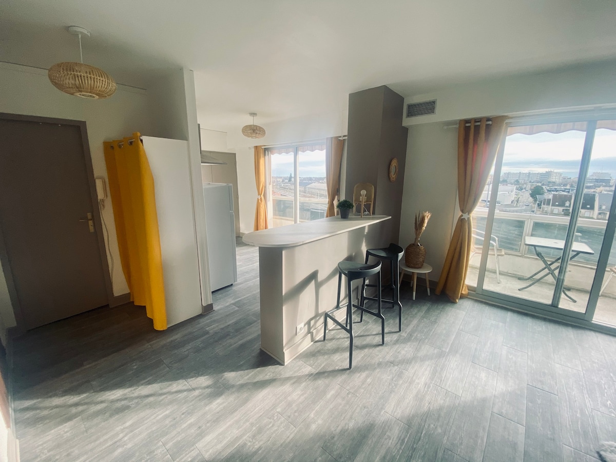 An open living space is observed, featuring a spacious kitchen island with seating for three. Large windows provide natural light and offer a view of a balcony with outdoor seating. Soft drapes frame both windows, while light-colored cabinetry adds a modern touch.