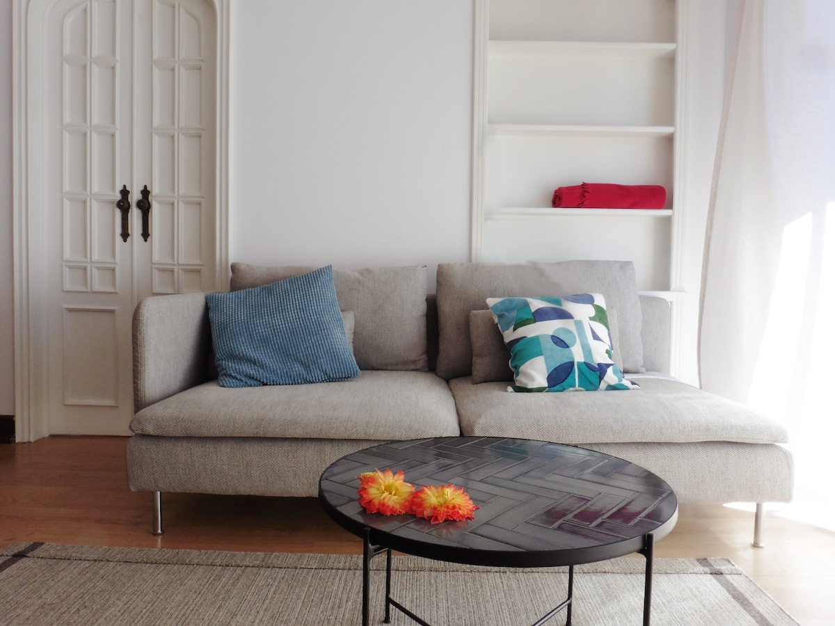 A light-filled living area displays a soft gray sofa adorned with patterned and solid-colored cushions. In front, a round black coffee table is accented with vibrant artificial flowers. A built-in shelf, partially visible, features a few items, contributing to a clean and organized space.