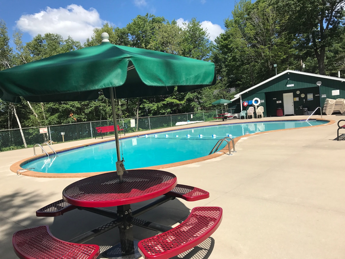 An inviting outdoor pool is surrounded by a concrete deck, featuring a shaded picnic table with red benches nearby. Lush greenery and trees provide a natural backdrop, while a clubhouse can be seen in the distance, adding to the tranquil atmosphere.
