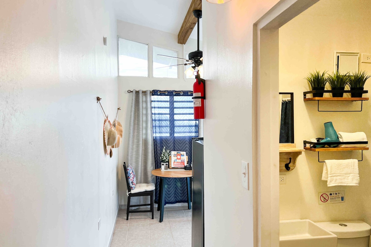 A glimpse into the apartment reveals a bright hallway leading to a cozy dining area with a round table and two chairs. Natural light filters through the nearby window, accentuated by soft curtains. A minimalistic bathroom area is visible, featuring neatly arranged toiletries and a towel rack.
