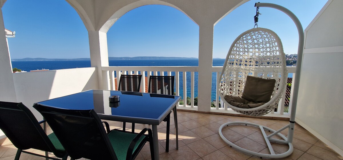 A private terrace features a glass-top dining table accompanied by four dark chairs. A hanging chair is visible in the corner, offering a relaxing space. The expansive sea view is framed by arched openings, with a clear blue sky and distant shoreline adding to the scenic backdrop.