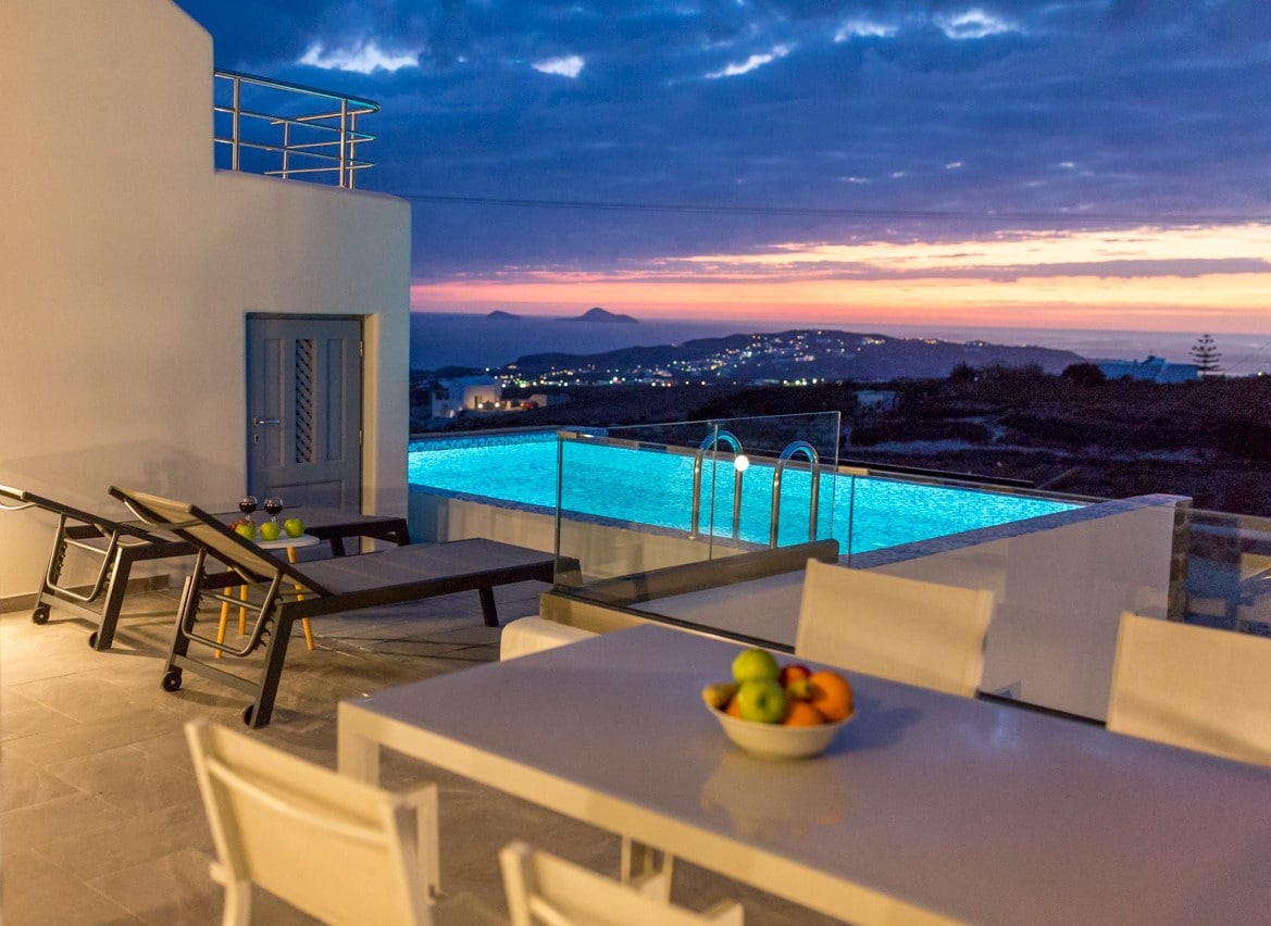 An outdoor dining area features a table with a fruit bowl, surrounded by white chairs. Sunbeds line the edge of a private swimming pool, which glows under a dusky sky, showcasing a view of the sunset and distant lights from the coastline.