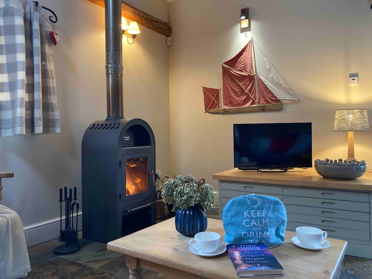A warm sitting area features a log burner at its center, providing a cozy focal point. Nearby, a wooden table holds two cups and a decorative blue item. A television is positioned against a wall, with nautical-themed decor enhancing the rustic ambiance.