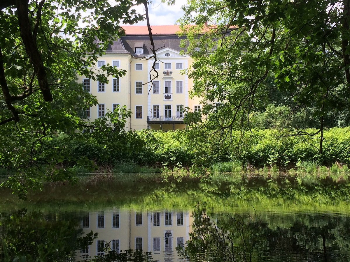 A historic building is reflected in the calm waters of a pond, framed by lush greenery and tree branches. The structure features multiple windows and a balcony, showcasing a yellow facade that contrasts with the surrounding nature.