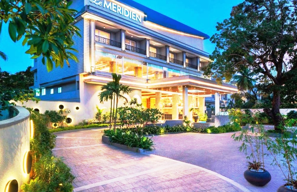 The exterior of Le Méridien features a contemporary design set against a twilight sky. Lush greenery surrounds the entrance, complemented by well-maintained pathways and garden areas. The building's large windows reflect soft lighting from within, creating an inviting atmosphere.