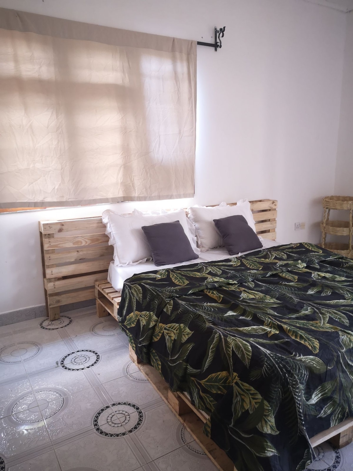 A spacious bed is presented with a dark leaf-patterned blanket and two grey cushions. Light filters in through a window covered by a beige curtain, illuminating the room's minimalist design, which features a tiled floor and wooden furniture.