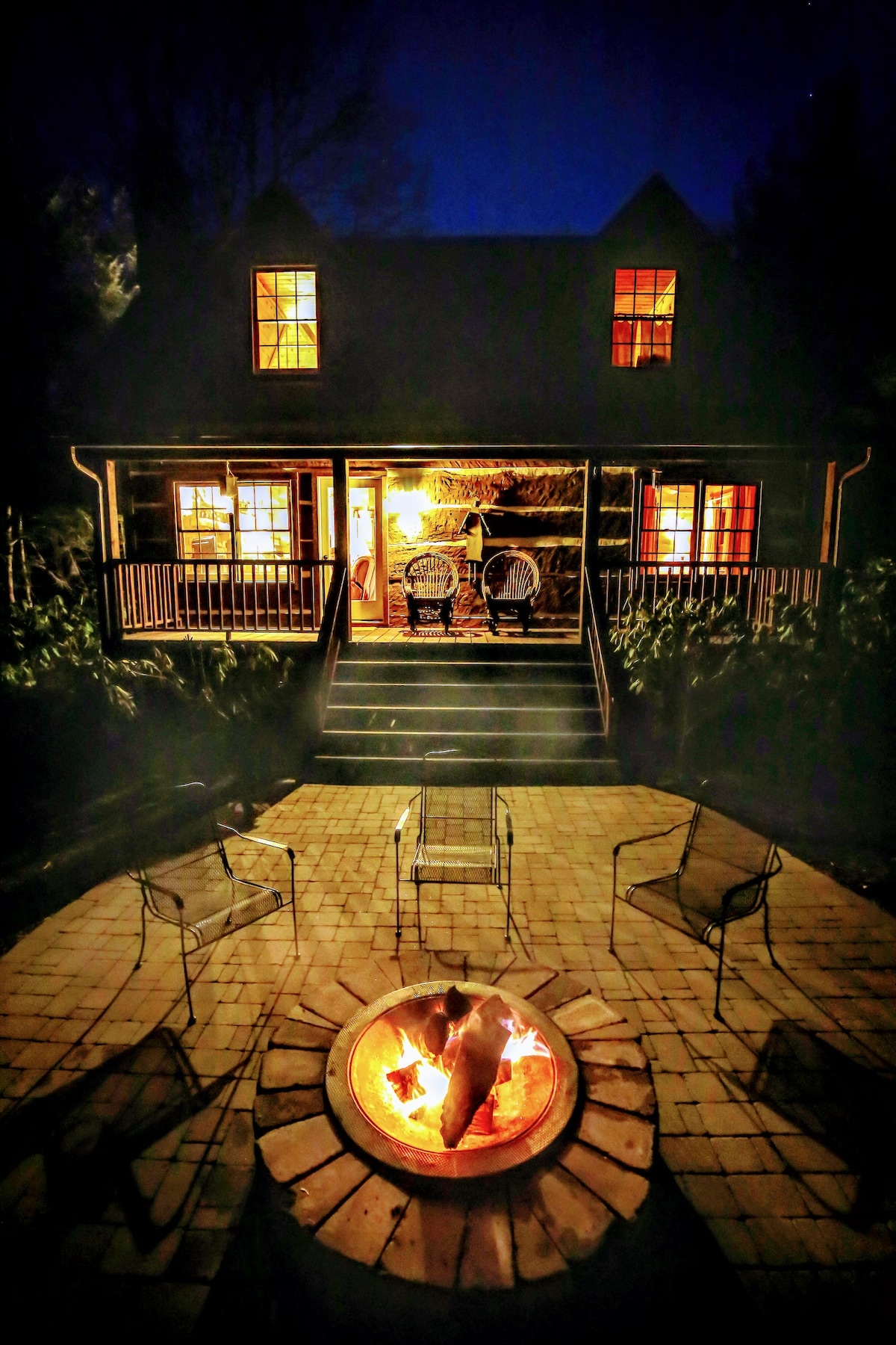 A cozy outdoor fire pit is surrounded by four metal chairs, inviting gatherings under the evening sky. The cabin's warm lights glow through the windows, while two rocking chairs are positioned on the covered porch, providing a peaceful view of the surrounding landscape.