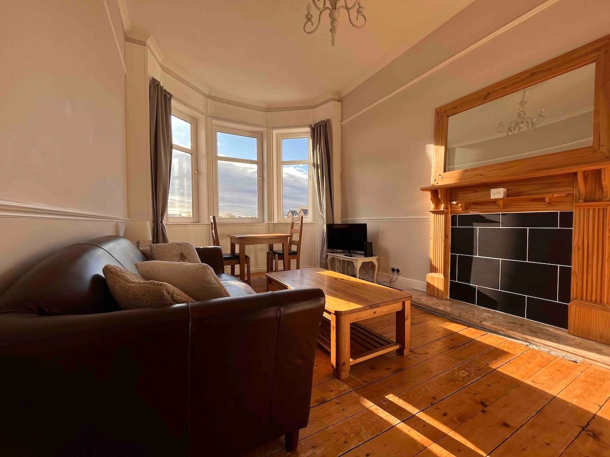 A bright lounge area features a bay window allowing ample natural light. Comfortable seating is arranged around a wooden coffee table, with a small dining table positioned nearby. A fireplace with a mirror above serves as a focal point, enhancing the room's cozy atmosphere.