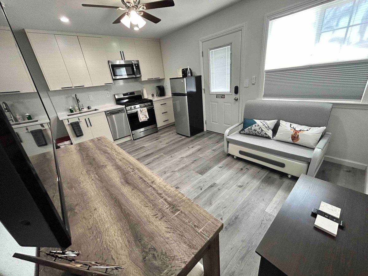 A modern kitchen is combined with a living area, featuring stainless steel appliances and a dining table. A comfortable couch with decorative pillows and a coffee table are included. Natural light enters through a window, illuminating the light-colored cabinetry and flooring.