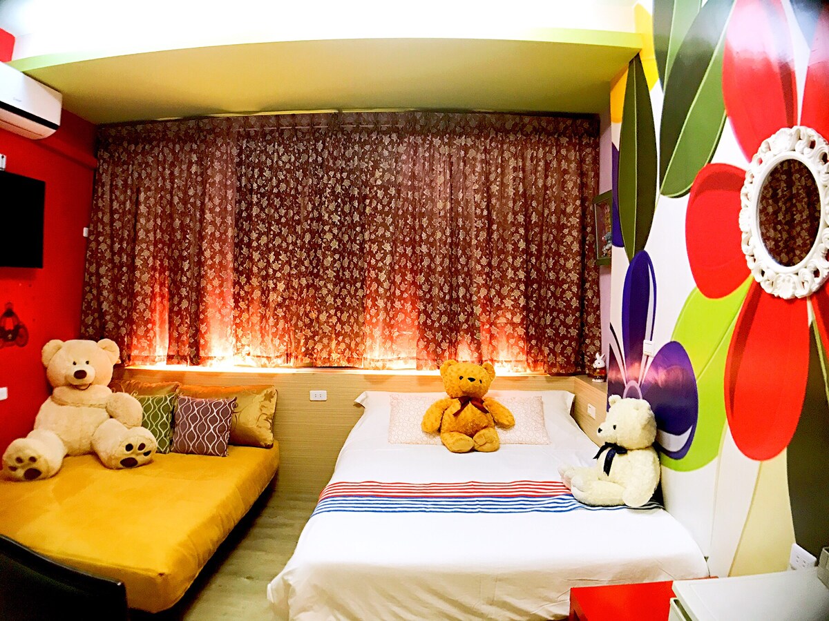 The room features a spacious bed dressed in white linen, complemented by colorful striped accents. A cozy seating area with a yellow sofa is adorned with plush teddy bears, enhancing the playful decor. The vibrant wall design adds a touch of whimsy to the space.