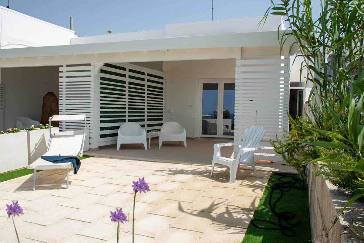 A spacious outdoor patio features white seating arrangements and a low green hedge. Sunlight illuminates the area, highlighting the stone flooring. A glimpse of the sea can be seen through the glass doors, adding a serene backdrop to the setting.