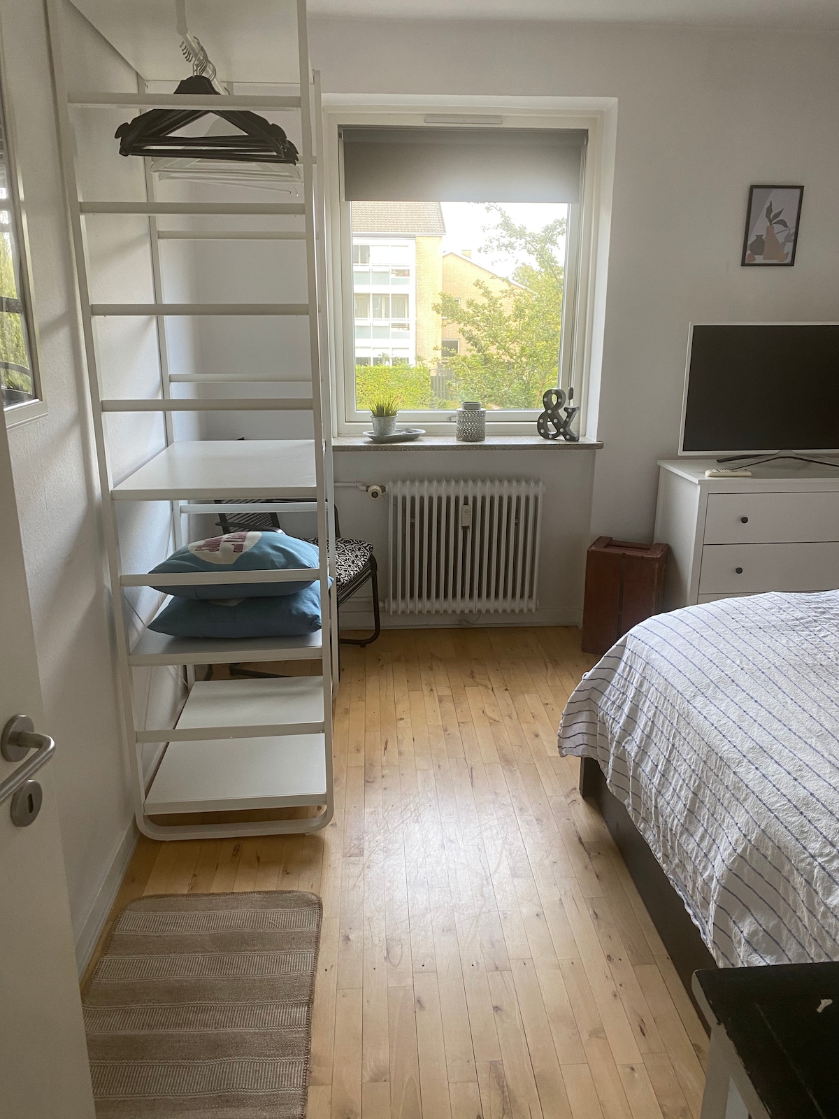 A spacious bedroom is visible, featuring a large window that allows natural light to fill the room. A bed with a checkered duvet is seen, along with a dresser and a television. A shelf holds decorative items and pillows, creating an organized space.