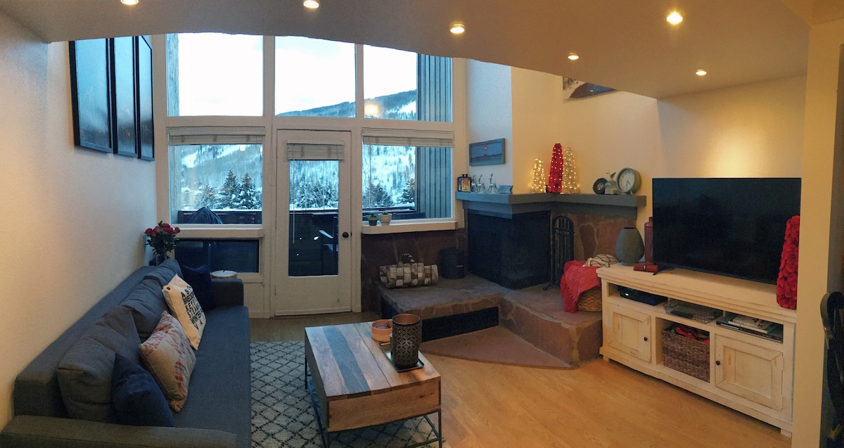 A spacious living area features floor-to-ceiling windows showcasing views of the Vail ski slopes. A comfortable sofa is arranged next to a wooden coffee table, with decorative items and a flat-screen television present. A stone fireplace adds warmth to the inviting space.